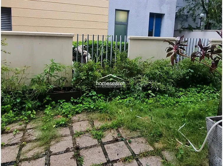 This outdoor area consists of a paved terrace or patio space surrounded by a low concrete wall and metal fencing. The area features overgrown vegetation and garden beds, suggesting it requires some landscaping maintenance to restore its curb appeal.