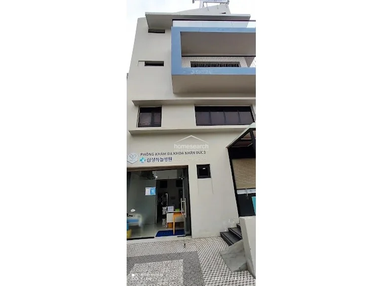 This image displays the exterior facade of a multi-story commercial building currently operating as a medical clinic. The structure features a modern, minimalist design with a prominent blue-accented balcony on the upper level and a glass-paneled entrance at street level. The exterior is finished in a clean, off-white paint, and the ground floor includes patterned tile paving and a small set of steps leading to a side entrance.