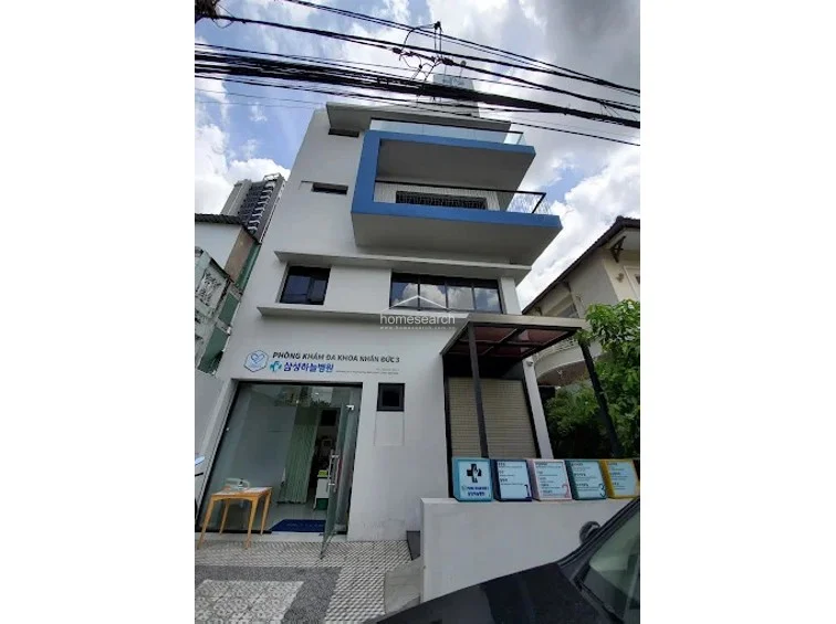 This wider angle captures the full vertical profile of the building, highlighting its three-story layout and the prominent signage for "Phòng Khám Đa Khoa Nhân Đức 3." The property includes a small, gated outdoor area with a series of informational placards and a covered entryway, suggesting a functional layout designed for patient access. The building appears to be in good condition, featuring contemporary architectural elements like the cantilevered balcony and large, dark-tinted windows that provide a professional aesthetic.