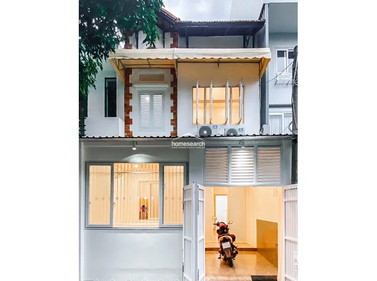 This two-story townhouse features a white facade with brick accents and a protective awning over the upper windows. The ground floor includes a gated entrance area suitable for motorcycle parking, with security bars on the windows and external AC units installed for the upper level.