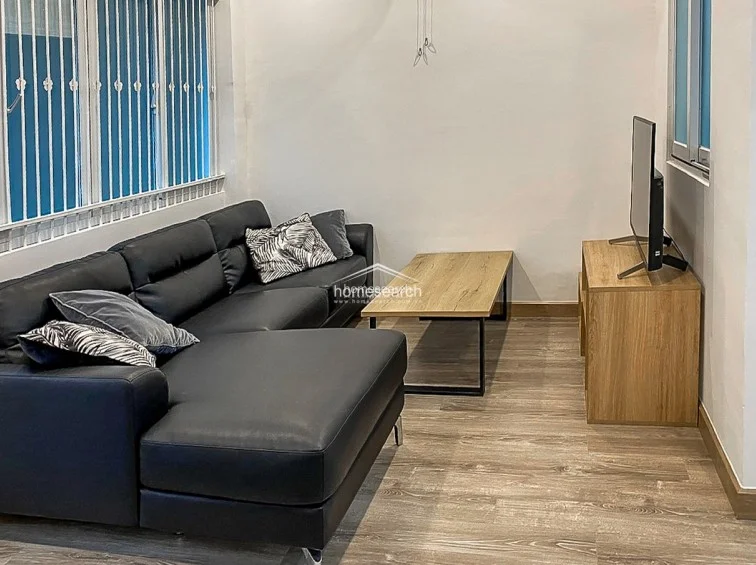 The living area is finished with wood-look flooring and neutral white walls, creating a clean and modern aesthetic. It is furnished with a black L-shaped sofa, a wooden coffee table, and a media console, while large windows with security bars allow for natural light.