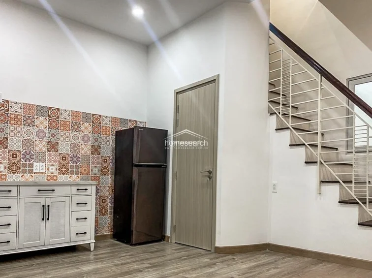This kitchen area features a decorative, multi-patterned tile backsplash that adds a vibrant touch to the otherwise minimalist white cabinetry. The space is efficiently laid out under a staircase with a dark-toned refrigerator and wood-look flooring, maintaining a tidy and well-maintained appearance.