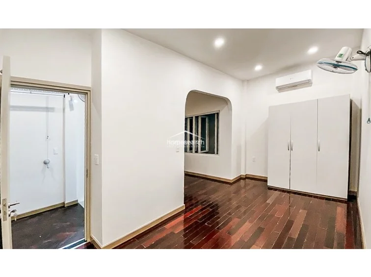 This bedroom features polished dark wood flooring and a large white wardrobe, providing ample storage space. The room includes a wall-mounted AC unit, a ceiling fan, and an arched doorway leading to an adjoining area, all illuminated by recessed ceiling lights.