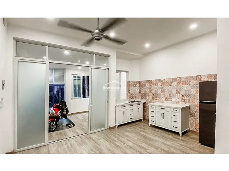 This open-concept kitchen and living space is separated from the entrance by large sliding glass doors, which help maintain privacy while allowing light to flow through. The kitchen is equipped with white cabinetry and a striking patterned backsplash, complemented by a modern ceiling fan and wood-tone flooring throughout.