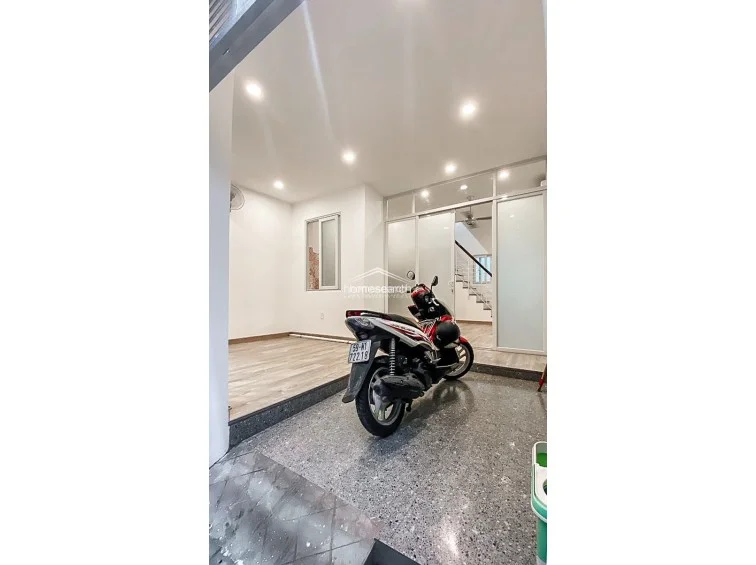 This ground-floor area serves as a versatile entrance or garage space, featuring durable speckled stone flooring and a raised section with light wood-look flooring. The space is brightened by recessed ceiling lights and connects to the interior via a modern glass-paneled partition, offering a clean and well-maintained transitional zone.