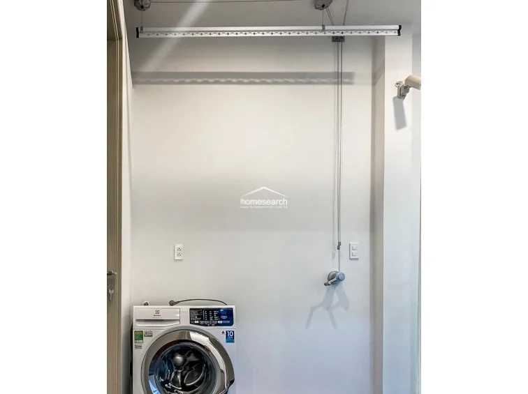 This dedicated laundry area is equipped with a modern Electrolux front-load washing machine and a convenient ceiling-mounted pulley drying rack. The space is finished with neutral white walls and includes a wall-mounted security camera, indicating a focus on utility and home safety.