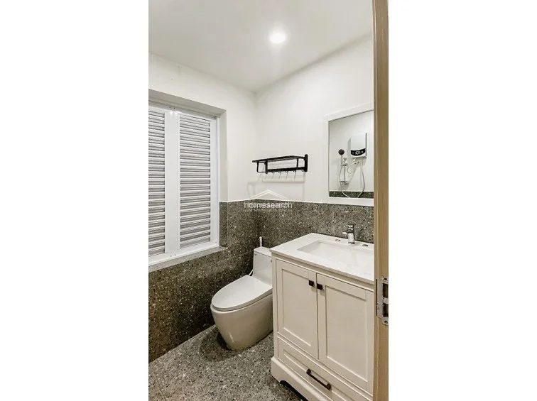 This compact, modern bathroom features a sleek white vanity cabinet with a marble-top sink and a contemporary toilet. The room is finished with high-contrast speckled stone flooring and matching wall tiling, complemented by a louvered window for ventilation and a wall-mounted water heater.