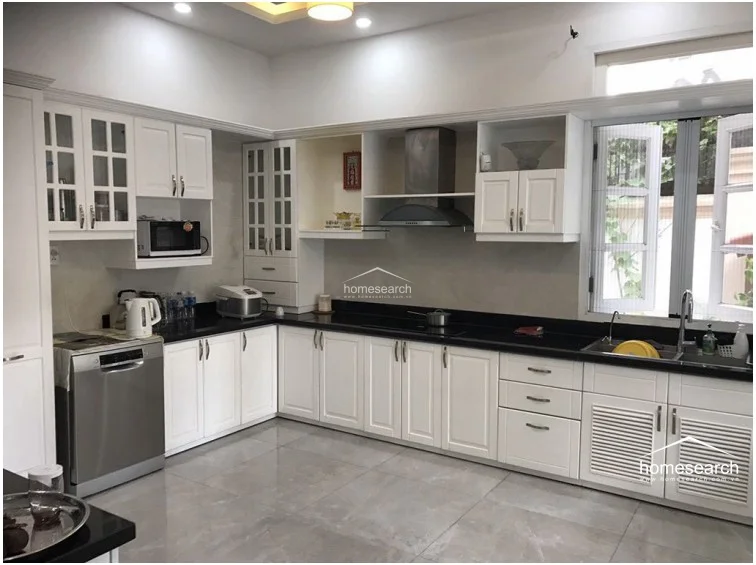 This spacious, L-shaped kitchen features crisp white cabinetry with glass-front accents and a sleek black countertop that provides a modern contrast. It is well-equipped with a stainless steel dishwasher, a built-in range hood, and large windows that invite ample natural light into the workspace. The room is finished with neutral, large-format floor tiles, maintaining a clean and well-kept aesthetic throughout.