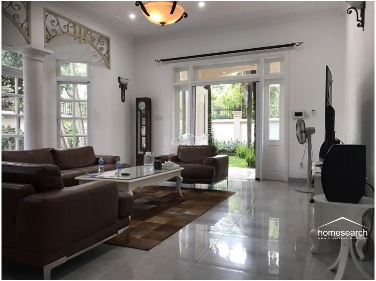The living room features an open-concept layout with polished light-colored floor tiles and a neutral color scheme that enhances the sense of space. Architectural details such as decorative archways and columns frame the room, which flows seamlessly toward the outdoor garden through large glass doors that provide ample natural light.