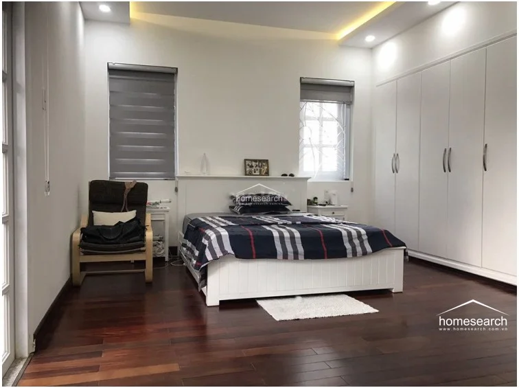 This primary bedroom offers a warm and inviting atmosphere, featuring rich dark wood flooring and a large, built-in white wardrobe for extensive storage. Natural light streams in through two windows fitted with modern roller blinds, highlighting the minimalist white walls and the comfortable bed setup. The space is well-maintained and includes a cozy reading chair, creating a peaceful retreat.