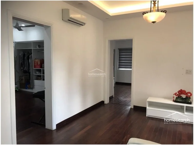 This transitional living area serves as a central hub, connecting to both a walk-in closet space and an adjacent room through elegant doorways. The room is finished with polished dark wood flooring and white walls, complemented by a sophisticated tray ceiling with recessed lighting and a decorative chandelier. A wall-mounted AC unit ensures climate control, while the overall condition reflects a high standard of maintenance.