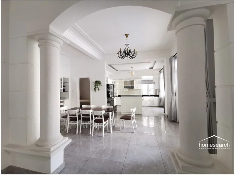 The dining and kitchen area is defined by grand white columns and an expansive archway, creating a sophisticated transition between zones. The space is bright and airy, featuring white cabinetry, high-gloss floor tiles, and a minimalist aesthetic that highlights the home's well-maintained, contemporary interior design.