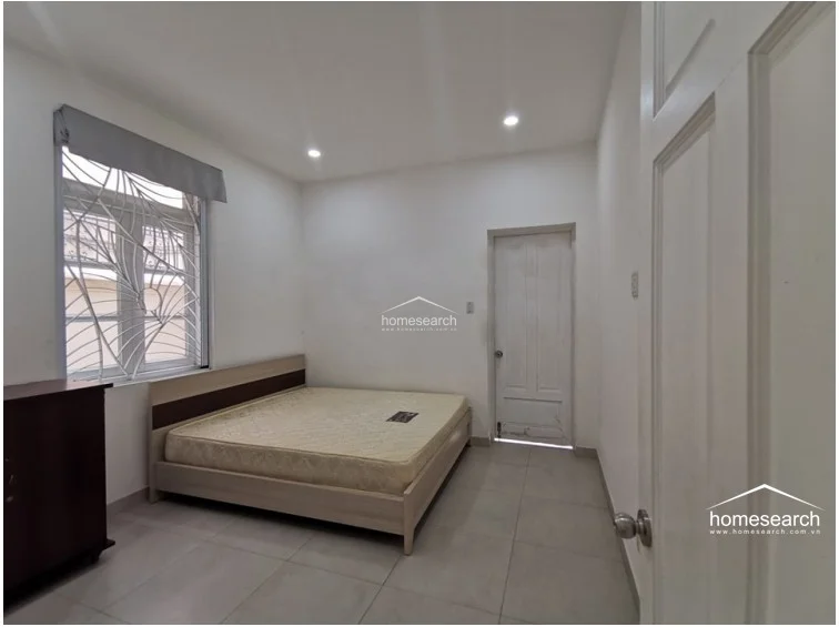 This secondary bedroom is functional and bright, featuring neutral large-format floor tiles and simple white walls that create an airy feel. It includes a basic bed frame and a window secured with decorative iron grilles, which allows for natural ventilation and light. The room is clean and well-maintained, offering a versatile space that could easily be adapted for guests or a home office.