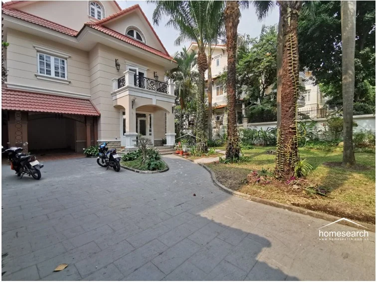 This exterior view showcases a multi-story villa with a classic architectural style, featuring a tiled roof, beige stucco walls, and a private driveway. The property includes a lush, well-maintained garden with mature palm trees and an integrated covered carport, reflecting a high level of maintenance and curb appeal.
