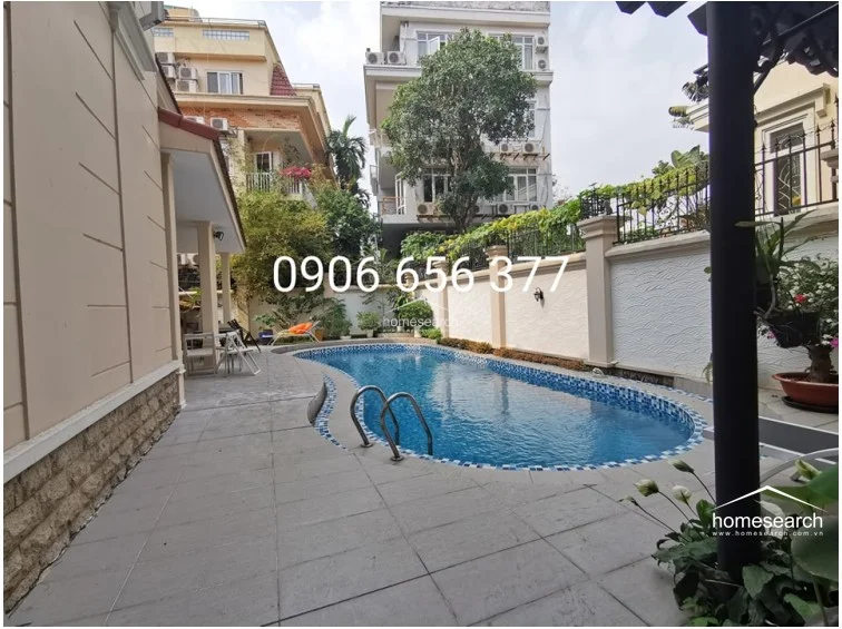 The outdoor area features a beautifully maintained kidney-shaped swimming pool surrounded by a spacious tiled deck, perfect for relaxation or entertaining. The space is enclosed by a high, decorative boundary wall and lush greenery, providing a private and tranquil oasis. The architecture of the home is visible, showcasing a well-kept exterior with modern finishes that harmonize with the garden setting.