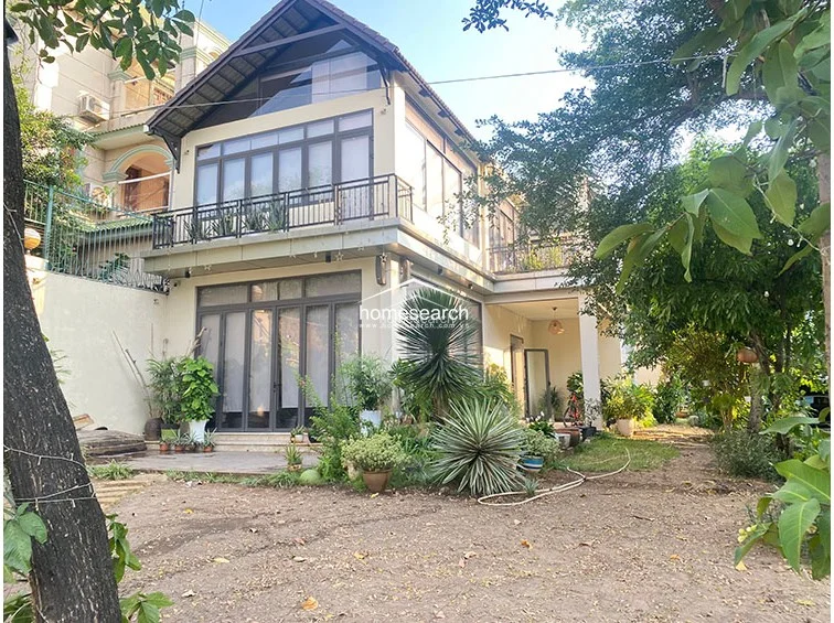 This two-story detached property features a modern, clean-lined architectural design with a prominent A-frame roof and extensive floor-to-ceiling glass windows. The exterior is surrounded by mature trees and lush greenery, including a ground-level patio and a spacious second-floor balcony with a metal railing. The structure appears to be in good condition, offering a seamless transition between indoor living areas and the natural outdoor environment.