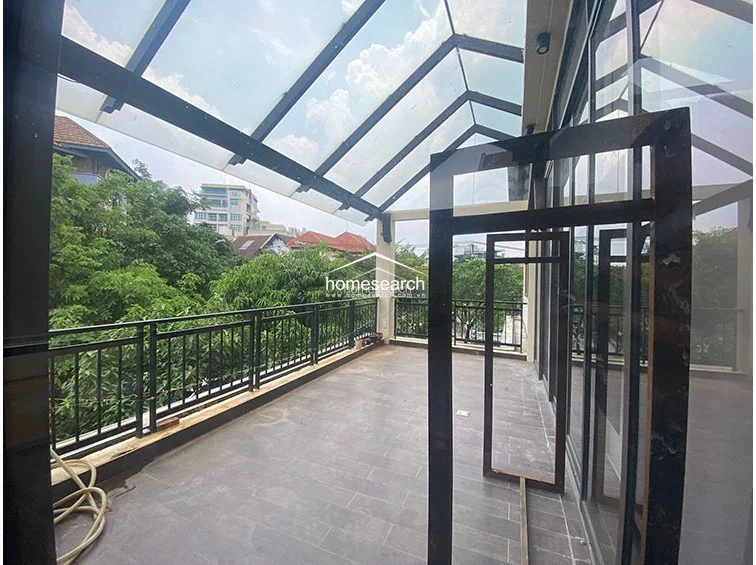 This spacious, covered terrace features a sleek glass-paneled roof and dark-toned floor tiles, providing a seamless transition between indoor and outdoor living. The area is enclosed by a black metal railing that offers an unobstructed, leafy view of the surrounding neighborhood, creating a bright and airy atmosphere. The structure appears well-maintained, with large glass sliding doors connecting the terrace directly to the interior of the property.