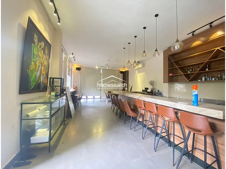 This commercial-style interior space functions as a bar or café, featuring a long, polished stone-topped service counter with leather bar stools. The room is illuminated by a series of stylish pendant lights and track lighting, with neutral wall finishes and large glass windows that provide ample natural light. The flooring consists of large-format light-colored tiles, and the space is accented by a large decorative painting and custom wooden shelving behind the bar.