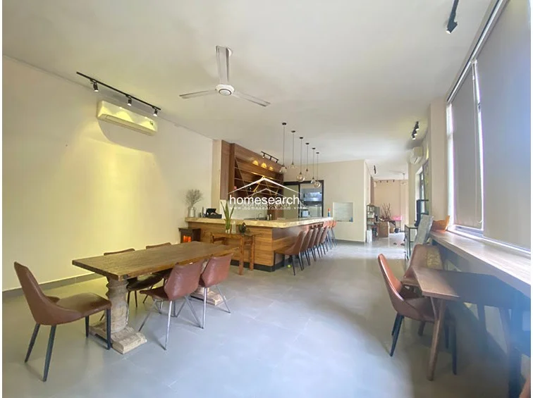 This dining area continues the open-concept flow of the property, showcasing a rustic wooden table paired with modern leather chairs. The room is equipped with a wall-mounted air conditioning unit and a ceiling fan, ensuring climate control throughout the space. Large windows with roller blinds allow for adjustable natural light, while the neutral color scheme and minimalist decor create a calm, professional atmosphere.