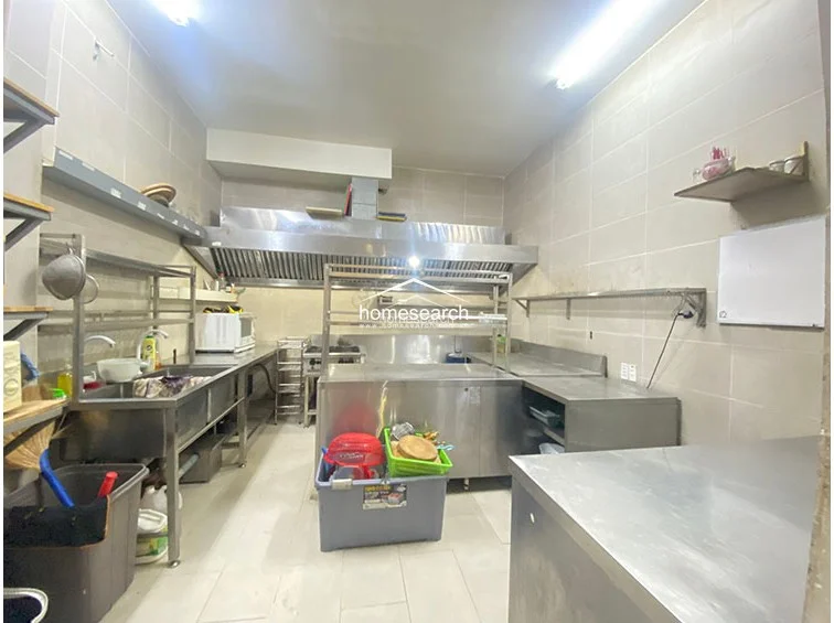 This professional-grade commercial kitchen is fully equipped with stainless steel surfaces, a large industrial exhaust hood, and deep sinks. The walls are finished with light-colored ceramic tiles, facilitating easy maintenance and high hygiene standards. The layout is optimized for high-volume food preparation, featuring ample storage shelving and efficient workspace flow.