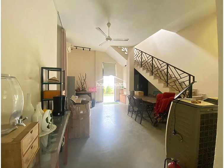 This multipurpose ground-floor area serves as a transition space, featuring an open staircase with a decorative black metal railing that leads to the upper level. The room is finished with neutral walls and tile flooring, providing a clean, functional space that connects the kitchen and service areas to the outdoor garden via a rear door. The area is currently used for storage and casual seating, maintaining a bright and airy feel due to the natural light entering from the rear entryway.