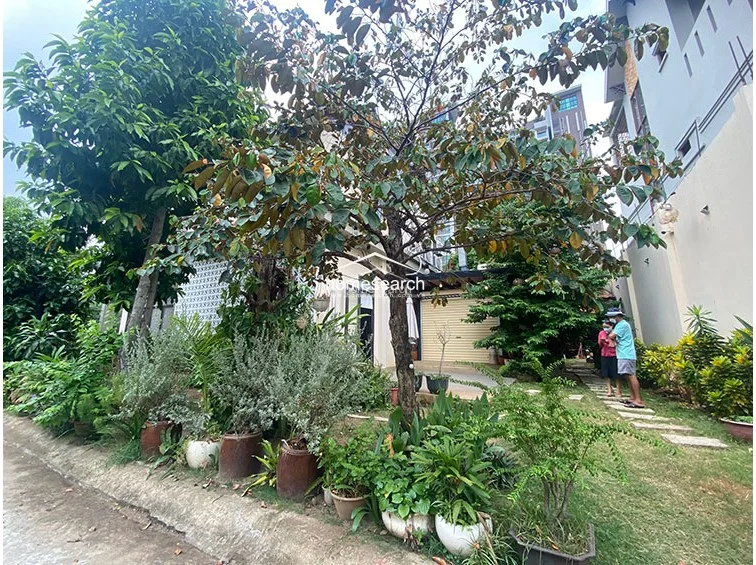This exterior view shows a lush, garden-like setting with a mature tree providing shade near the building entrance. The property features a paved walkway and a neutral-toned facade, suggesting a well-maintained, semi-private outdoor space.