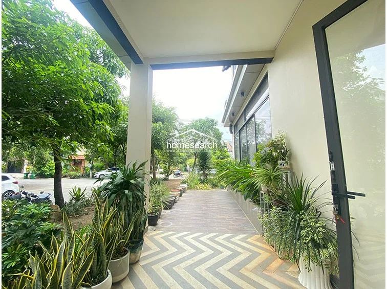 The entrance features a covered porch with a distinctive chevron-patterned tiled floor that transitions into wood-look tiles. Large glass doors and floor-to-ceiling windows provide excellent natural light and a seamless connection to the surrounding greenery.