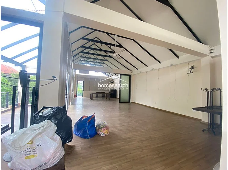 This expansive interior space features high, vaulted ceilings with exposed black structural beams, creating an airy and industrial-chic atmosphere. The room is finished with light wood-look flooring and neutral walls, currently configured as an open-plan area with a direct flow to an outdoor balcony.
