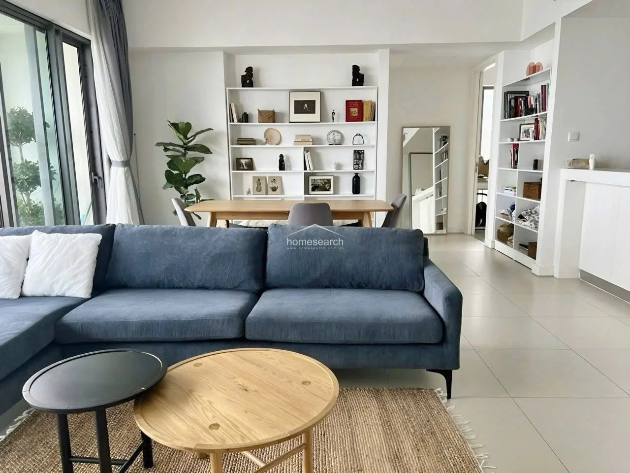 The living area is styled with a cozy, contemporary feel, featuring a plush blue sectional sofa, a textured jute rug, and a nested coffee table set. A built-in white bookshelf provides ample storage and display space, while a large mirror adds depth to the room. The space is well-maintained and bright, benefiting from the natural light streaming through the adjacent balcony doors.