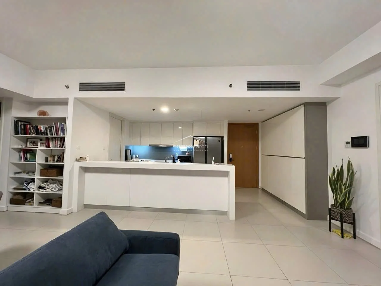 The kitchen and entryway area showcases a highly efficient, open-plan design with a large breakfast bar that serves as a visual divider. The space includes integrated ceiling-mounted AC units and extensive built-in storage, including a floor-to-ceiling cabinet unit near the entrance. The minimalist white cabinetry and light tile flooring contribute to the apartment's sleek, modern, and well-maintained appearance.