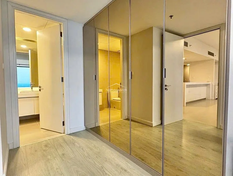 This transitional hallway area serves as a functional storage space, featuring a large, floor-to-ceiling mirrored wardrobe that reflects the adjacent bathroom and living area. The layout provides seamless flow between the private sleeping quarters and the common living spaces, with light-toned wood flooring and a bright, contemporary finish.