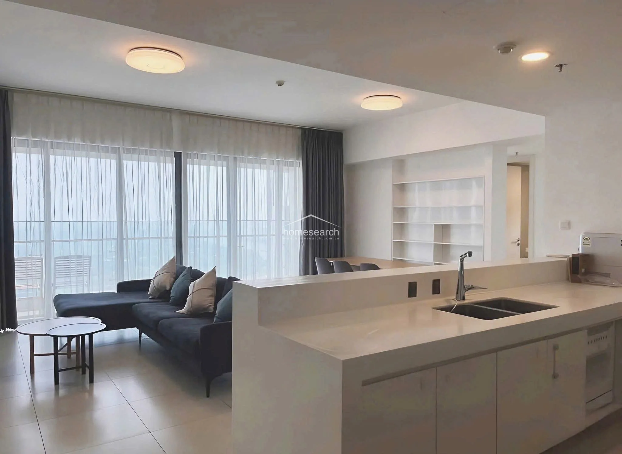 This open-concept living and dining area features a modern, minimalist aesthetic with neutral white walls and large-format light-colored floor tiles. The space is anchored by a large, dark blue sectional sofa and a breakfast bar that connects the kitchen to the living area, creating a seamless flow. Floor-to-ceiling windows with sheer and heavy curtains provide significant natural light and access to a balcony with an outdoor view.