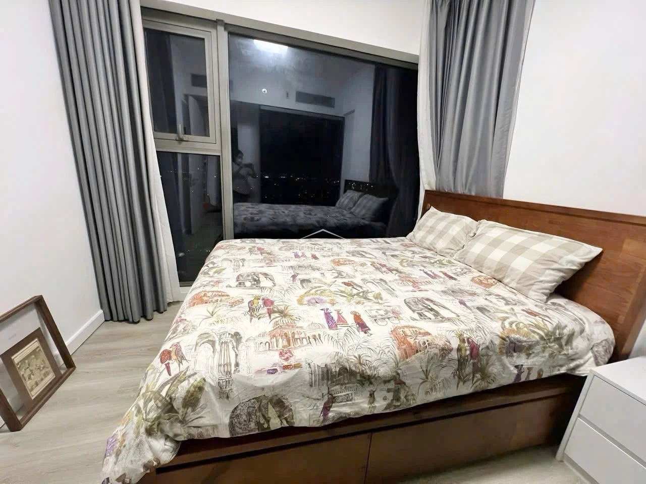 This bedroom features light wood-look flooring and a large floor-to-ceiling window that offers expansive night views and ample natural light. The room is furnished with a wooden bed frame and neutral-toned curtains, creating a clean, modern aesthetic with a well-maintained, minimalist feel.