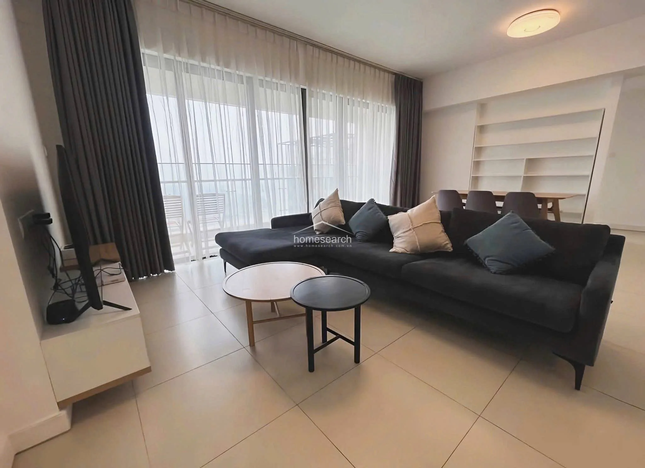 This view highlights the functional layout of the living room, emphasizing the connection between the seating area and the dining space. The room is kept bright and airy with a neutral color palette, complemented by the dark curtains and sofa that provide a sophisticated contrast. The overall condition is excellent, reflecting a modern, clean, and well-organized living environment.