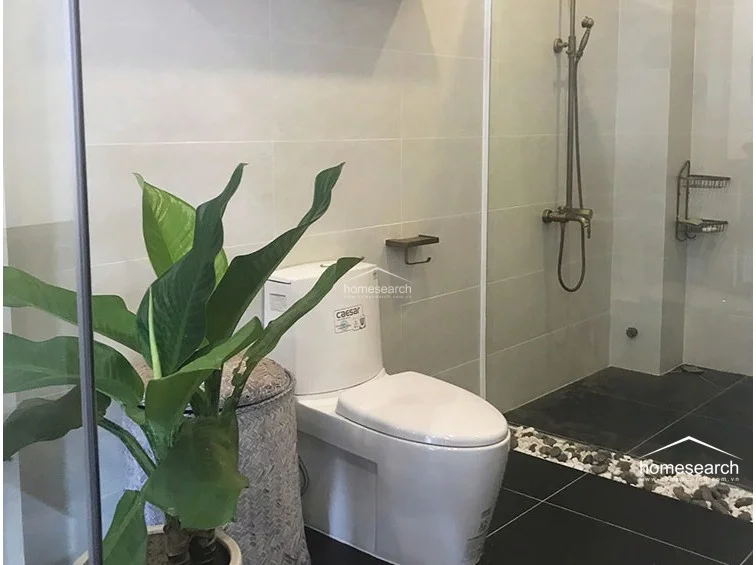 This bathroom features a modern, minimalist design with light-colored wall tiles and a contrasting black tiled floor. It includes a Caesar brand toilet, a wall-mounted shower fixture with a metallic finish, and a decorative pebble border that adds a natural, spa-like aesthetic.
