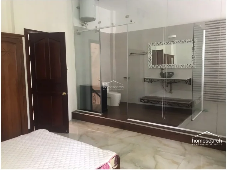 This bedroom features a unique, open-concept ensuite bathroom enclosed by floor-to-ceiling glass walls, creating a seamless flow between the sleeping and bathing areas. The room is furnished with a wooden wardrobe and a bed, while the bathroom area is elevated on a dark wood-look platform and includes a stylish vessel sink with a decorative mirror.