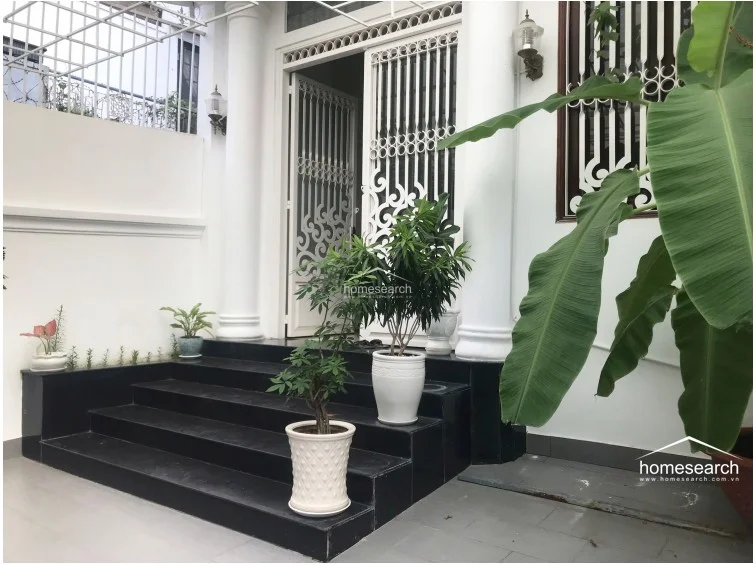 This shot captures the grand entrance steps leading to the home, flanked by classic white columns and lush potted greenery. The black stone steps provide a striking contrast against the white exterior walls and intricate metal security door, emphasizing a well-maintained and welcoming architectural style.