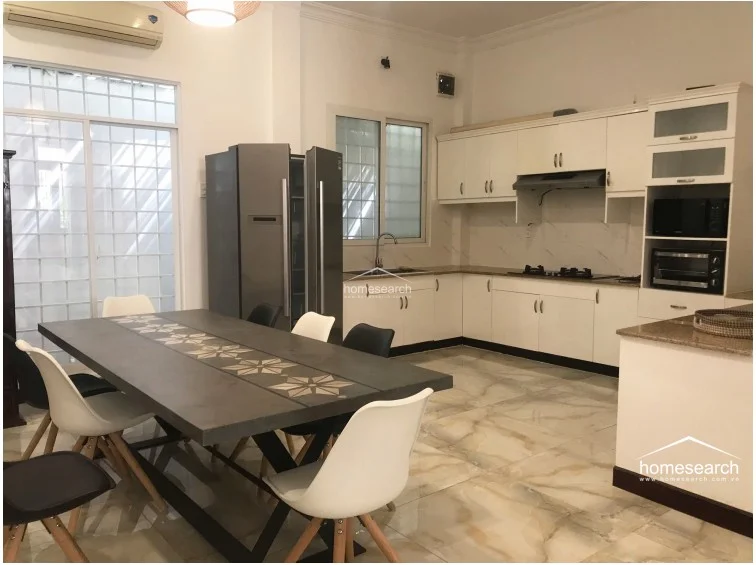 The kitchen is designed with an L-shaped layout featuring cream-colored cabinetry, granite countertops, and a full-height stainless steel refrigerator. It includes a dedicated dining area with a modern dark-topped table, large sliding glass doors with security grilles for natural light, and high-gloss marble-effect floor tiles.