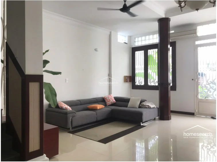 This open-concept living area is anchored by a large, modern grey sectional sofa and features polished light-toned flooring. The room is accented by a prominent dark wood structural column and a ceiling fan, with white walls that provide a clean, neutral canvas.