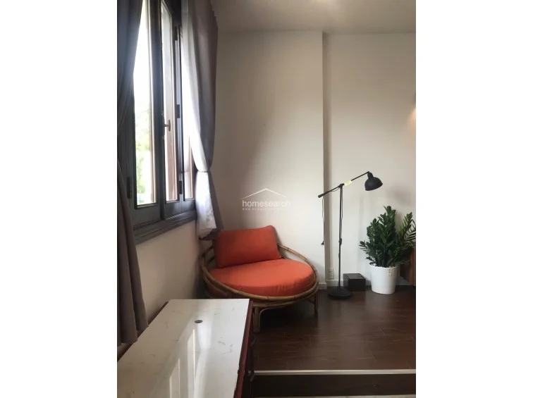 This intimate corner of the room serves as a cozy reading or relaxation nook, featuring a circular rattan chair with a vibrant orange cushion. It is situated on a raised dark wood-look platform that contrasts with the main marble flooring, illuminated by a modern floor lamp and natural light from the adjacent window.