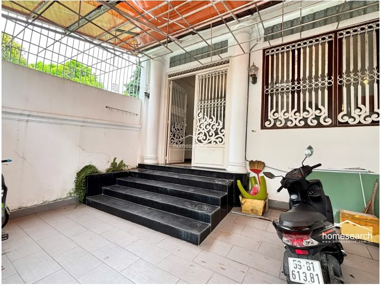This image shows the exterior entrance area of the property, which serves as a sheltered parking space for motorcycles and bicycles. The space is defined by an arched doorway, decorative metal security gates, and a raised staircase leading to the main entrance, all protected by a metal-framed overhead canopy.