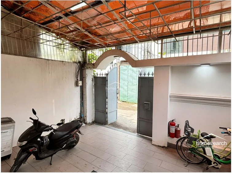This view highlights the covered entrance courtyard, which functions as a secure parking area for motorbikes and includes a washing machine tucked into the corner. The space is enclosed by a high metal gate and a protective overhead grid, providing a functional, semi-outdoor transition zone between the street and the home.