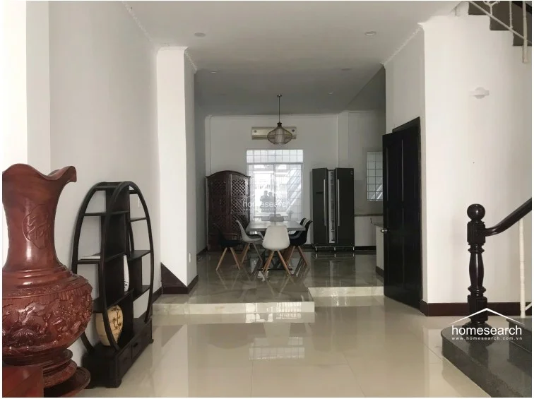 This open-plan living and dining area features a split-level design with polished cream tile flooring and crisp white walls. The space is anchored by a wooden staircase with a dark banister and includes a dining set illuminated by a modern pendant light, with a large refrigerator and air conditioning unit visible in the background.