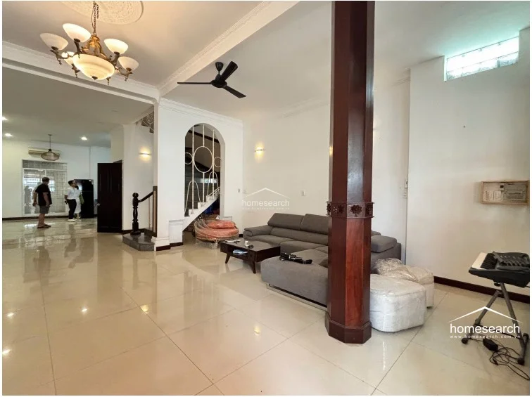 This spacious open-plan living area features high ceilings with decorative crown molding, a large central wooden support column, and polished light-colored tile flooring. The room flows seamlessly toward a rear kitchen area and includes a distinctive arched staircase enclosure, with white walls providing a bright, neutral backdrop for the modern gray sectional sofa.