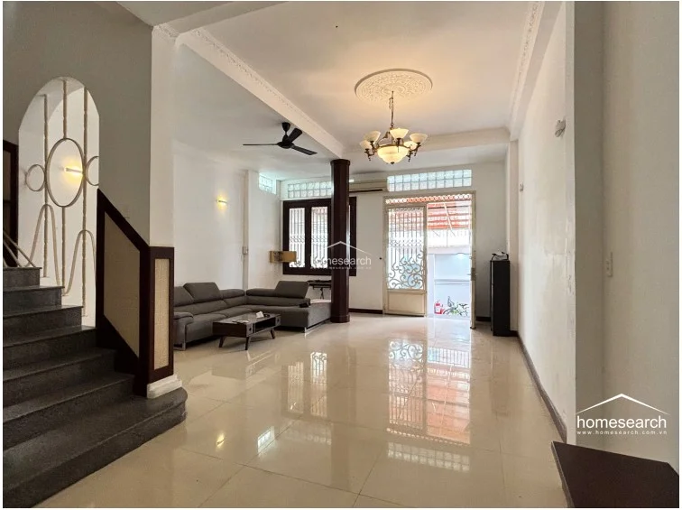 This view highlights the living room's depth, showcasing the transition from the front entrance area to the rear kitchen and staircase. The space is well-maintained, featuring a classic chandelier, a ceiling fan, and a consistent white color scheme that emphasizes the open, airy architectural design.