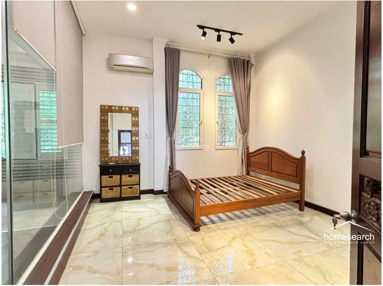 This bedroom features a unique open-concept layout with a glass-enclosed ensuite bathroom, creating a modern and airy flow. The room is finished with light-veined marble flooring, white walls, and arched windows that provide ample natural light, complemented by a wall-mounted AC unit and a wooden bed frame.