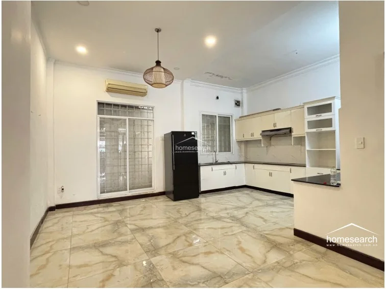 This kitchen and dining space is characterized by its clean, minimalist aesthetic, highlighted by a contemporary woven pendant light and large-format marble-patterned floor tiles. The area is equipped with a wall-mounted air conditioning unit and provides ample storage through extensive upper and lower cabinetry, maintaining a bright and functional environment.