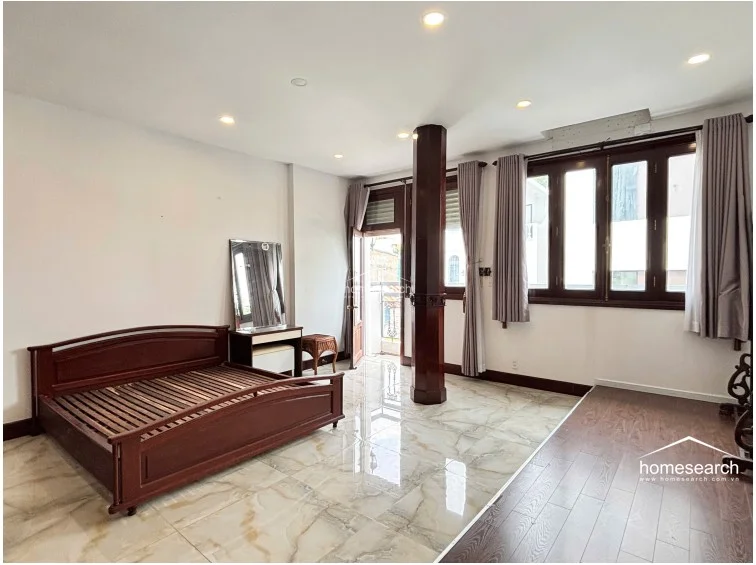 This spacious bedroom is anchored by a prominent, dark-stained wooden support column that adds an architectural focal point to the room. The layout includes a combination of marble and wood-look flooring, a wooden bed frame, and direct access to a small balcony, all under a ceiling with recessed lighting.