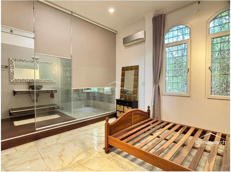This view highlights the bedroom's ensuite bathroom, which is separated from the sleeping area by a glass partition that can be obscured by a roller blind for privacy. The bathroom features a stylish vessel sink on a floating shelf and a vanity mirror, while the bedroom itself remains bright and minimalist with its marble floors and arched windows.
