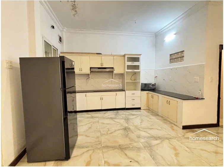This spacious kitchen features cream-colored cabinetry with a black countertop and large, light-toned marble-effect floor tiles. The layout is open and functional, with a large refrigerator and a dedicated space for a microwave, illuminated by a bright ceiling light fixture.